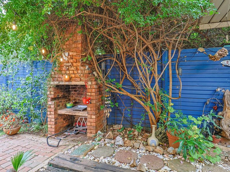 Outdoor patio area featuring a built-in brick braai, brick paving, and a blue timber fence, surrounded by established garden greenery.