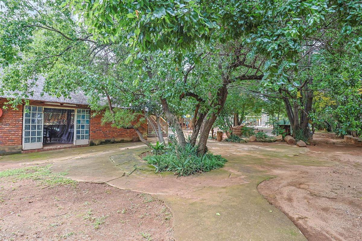 A spacious rear garden area featuring mature trees, a paved patio, and an exposed brick building with a thatched roof.