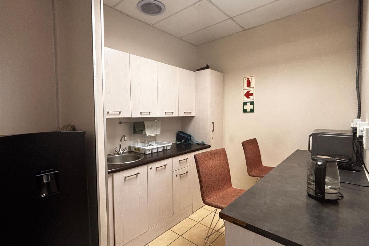 A functional office kitchenette featuring light wood-finish cabinetry, dark laminate countertops, a stainless steel sink, and tiled flooring.