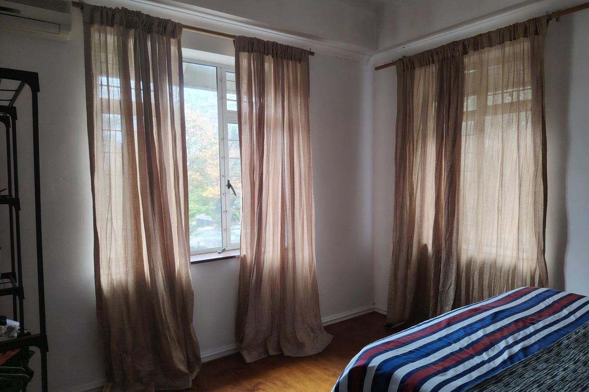 A bedroom featuring hardwood flooring, large windows with curtain rods, and a wall-mounted air conditioning unit.