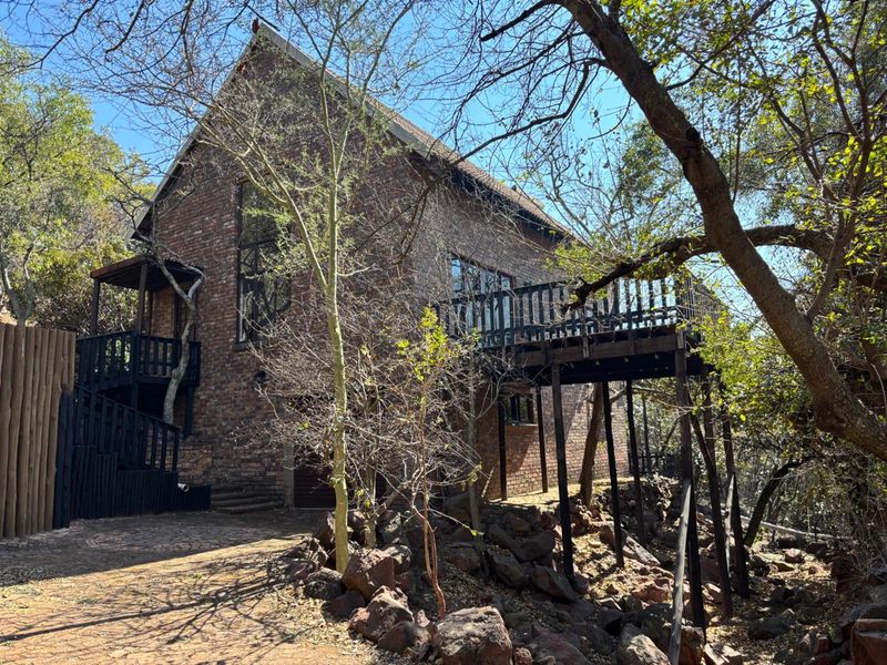A rustic brick house featuring a raised wooden deck and balcony, set within a natural, wooded landscape with a paved driveway.