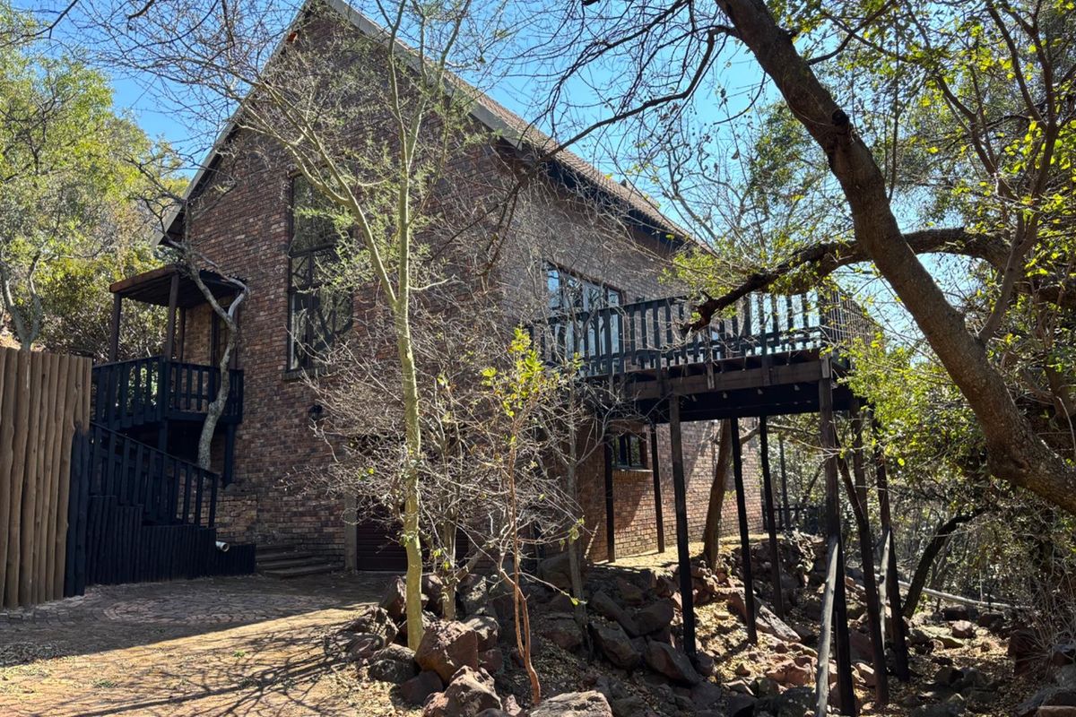 A rustic brick house featuring a raised wooden deck and balcony, set within a natural, wooded landscape with a paved driveway.