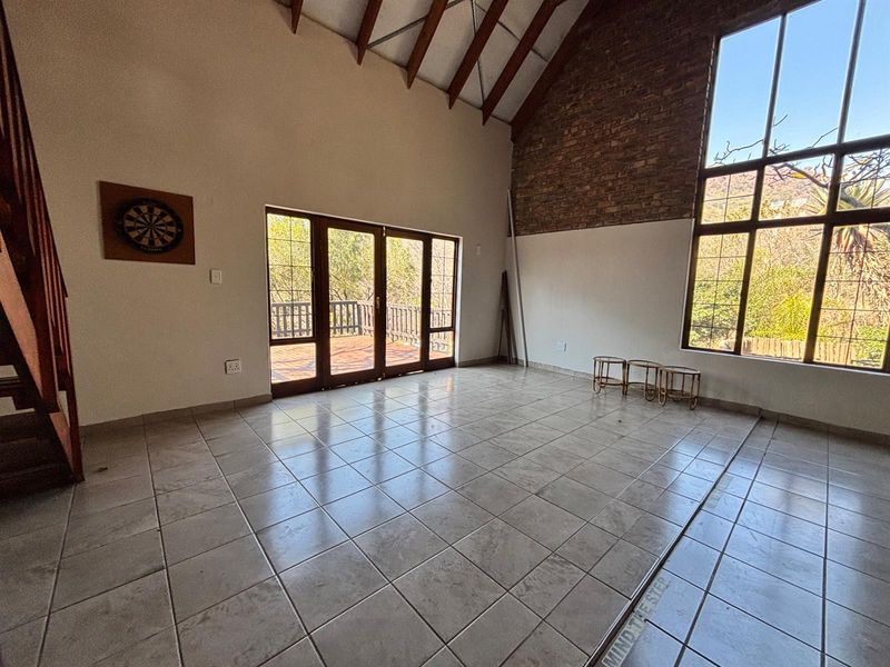Spacious living area featuring a vaulted ceiling with exposed timber rafters, a brick feature wall, tiled floors, and large windows leading to a deck.