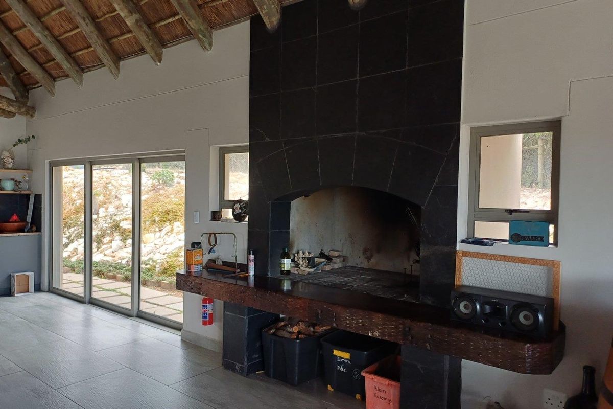 Spacious living area featuring a large built-in indoor braai with a dark tiled surround, exposed thatch roof beams, and sliding glass doors to the garden.