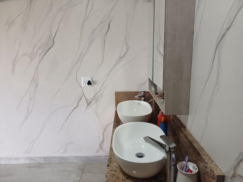 A modern bathroom vanity featuring dual vessel sinks, granite countertop, and a wall-mounted mirrored medicine cabinet against marble-effect wall cladding.