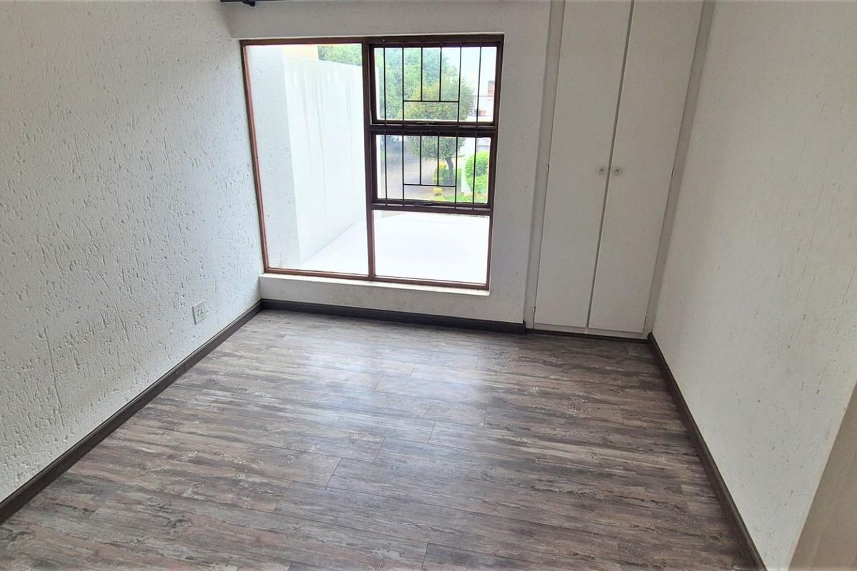 A bright, empty bedroom featuring modern laminate flooring, built-in cupboards, and a large window with security bars.