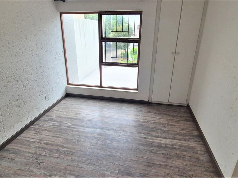 A bright, empty bedroom featuring modern laminate flooring, built-in cupboards, and a large window with security bars.