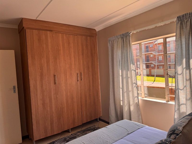 A well-maintained bedroom featuring a large built-in wooden wardrobe, tiled flooring, and a window with security bars overlooking the complex grounds.