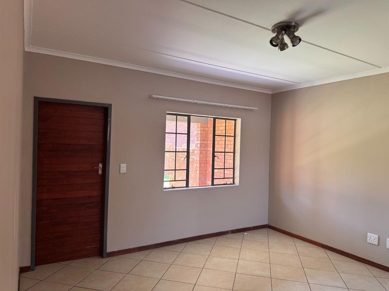 A clean, empty bedroom featuring neutral wall tones, tiled flooring, a solid wooden door, and secure window burglar bars.