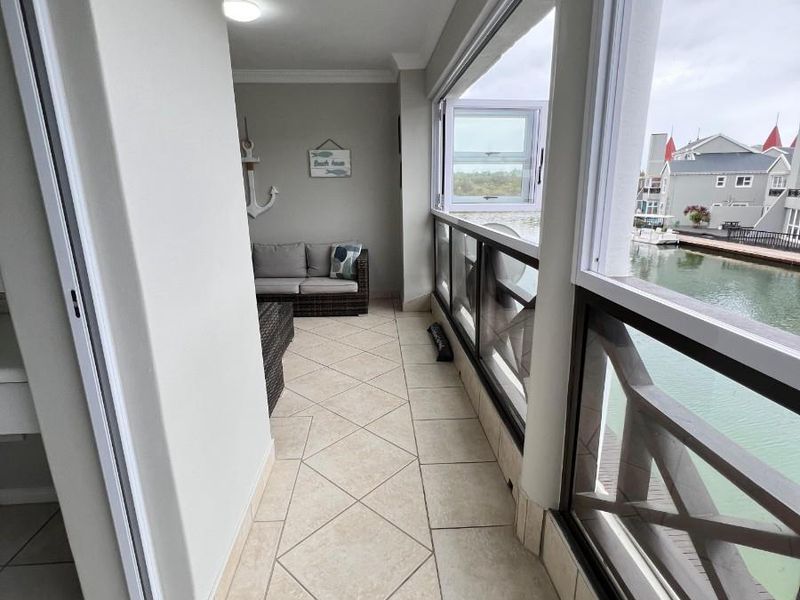 A covered balcony featuring tiled flooring, a glass balustrade, and sliding windows, offering a scenic view of the adjacent waterway.