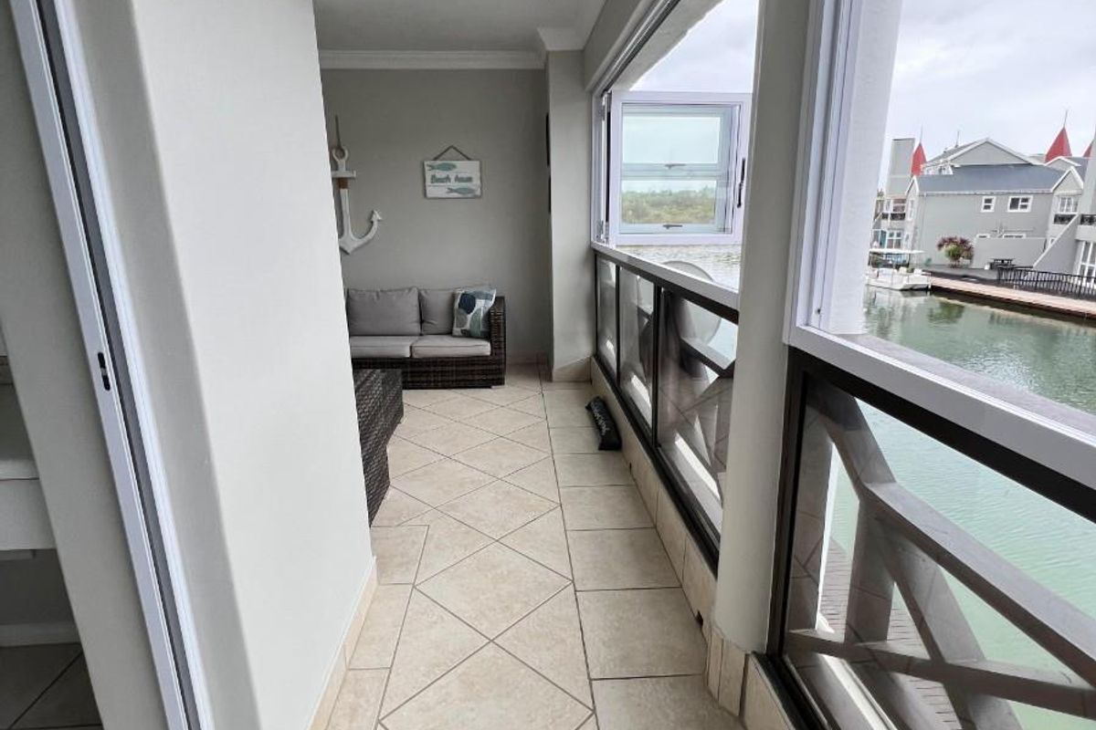 A covered balcony featuring tiled flooring, a glass balustrade, and sliding windows, offering a scenic view of the adjacent waterway.