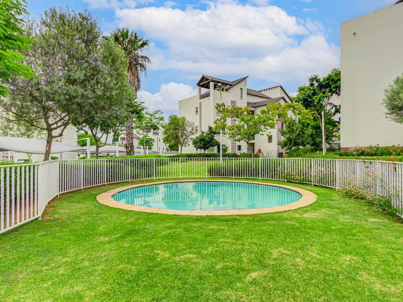 A well-maintained communal swimming pool area surrounded by manicured lawns and secure metal perimeter fencing within the apartment complex grounds.