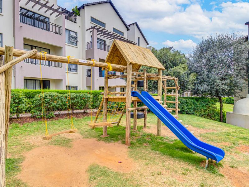 Communal garden area featuring a wooden playground set, slide, and a built-in outdoor fireplace set against the apartment complex facade.