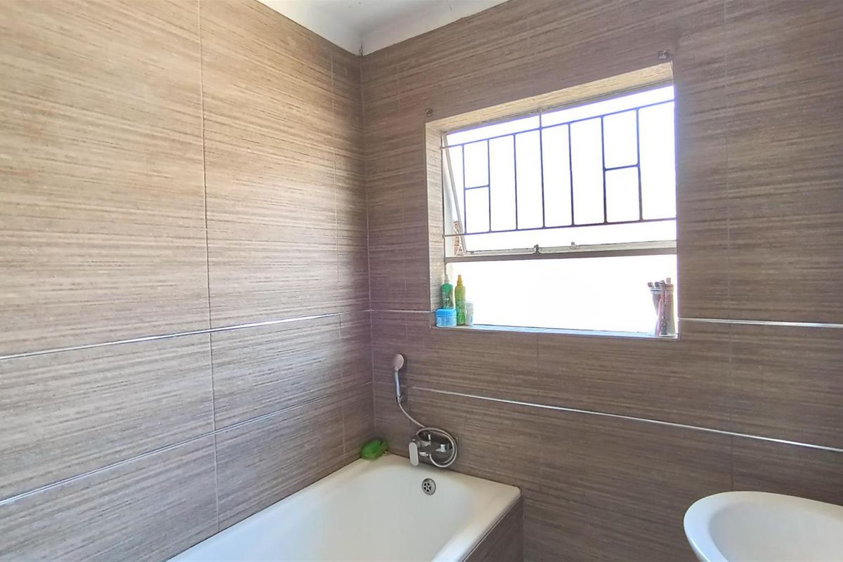 A functional bathroom featuring full-height wood-look wall tiling, a built-in bathtub with shower mixer, and a window with security bars.