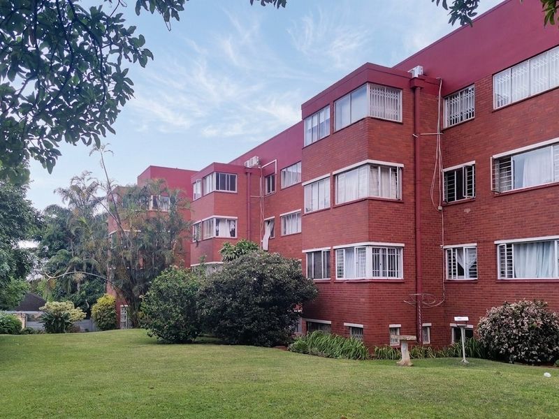 Rear view of a multi-story brick apartment building featuring large windows with security bars, set against a well-maintained communal garden area.