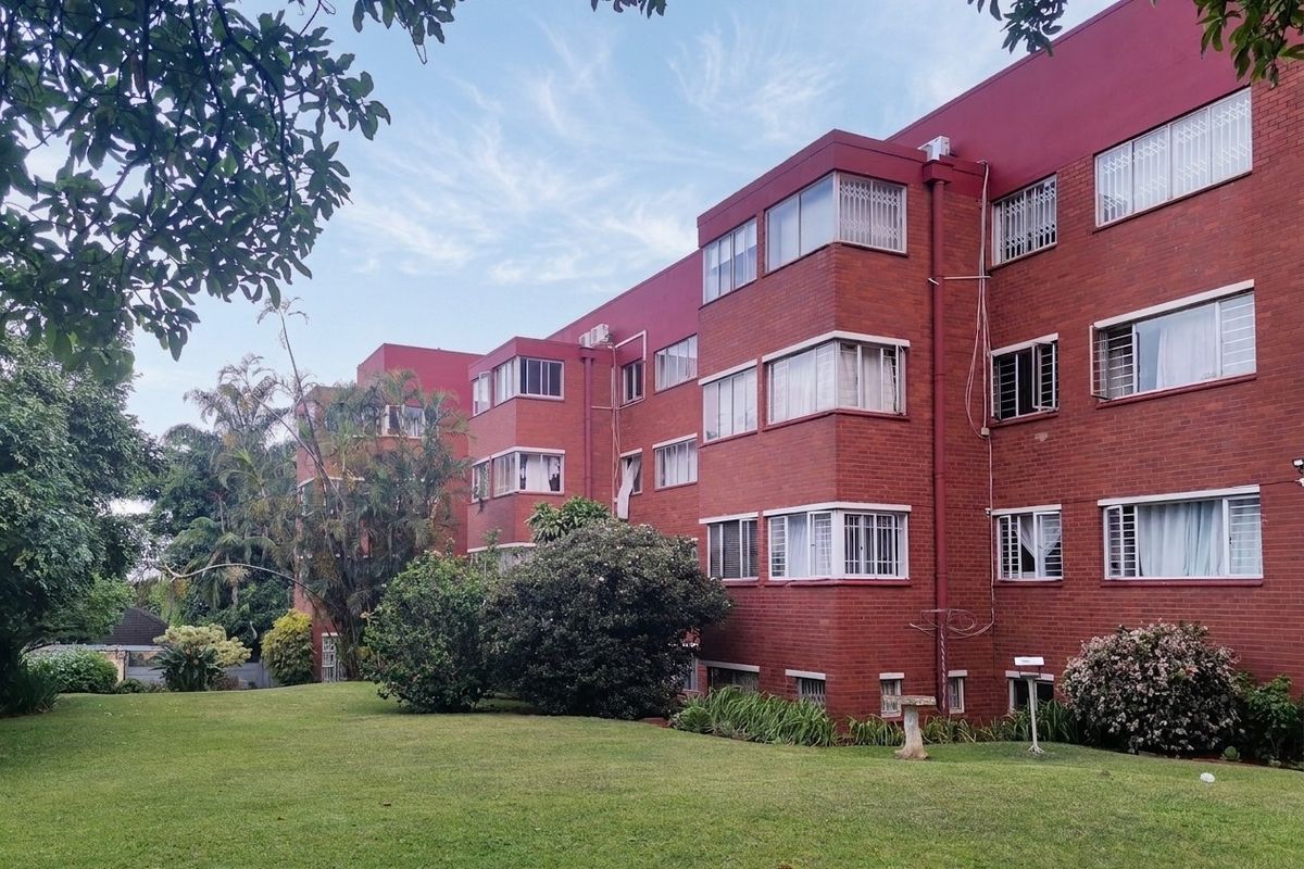 Rear view of a multi-story brick apartment building featuring large windows with security bars, set against a well-maintained communal garden area.