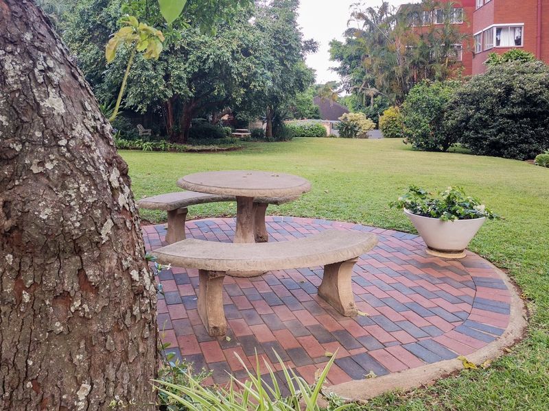 A well-maintained communal garden area featuring a circular brick-paved patio with integrated concrete seating and table, surrounded by established trees and lawn.