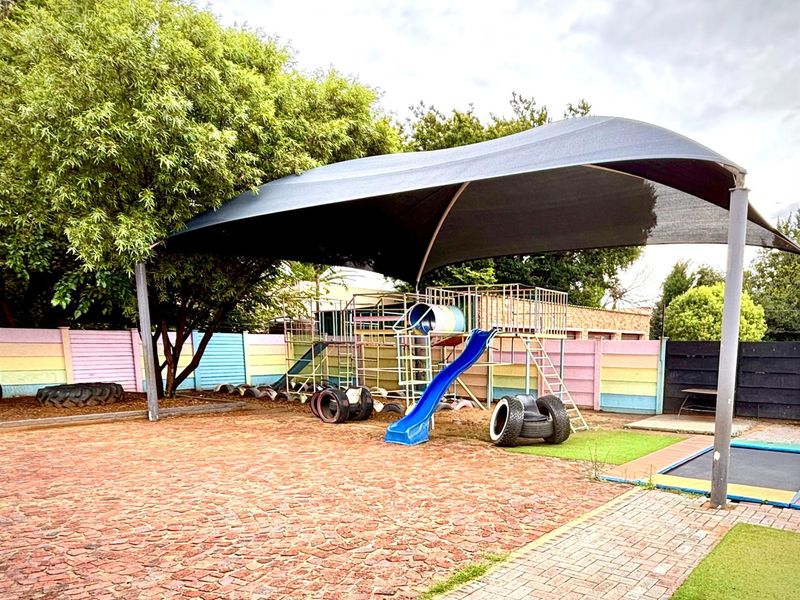 Outdoor play area featuring a large shade sail, paved ground, and fixed playground equipment within a secure perimeter wall.