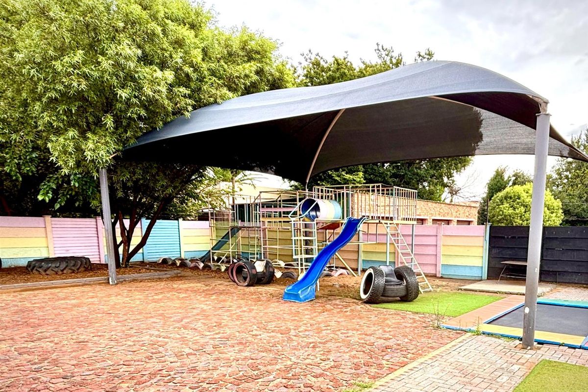Outdoor play area featuring a large shade sail, paved ground, and fixed playground equipment within a secure perimeter wall.