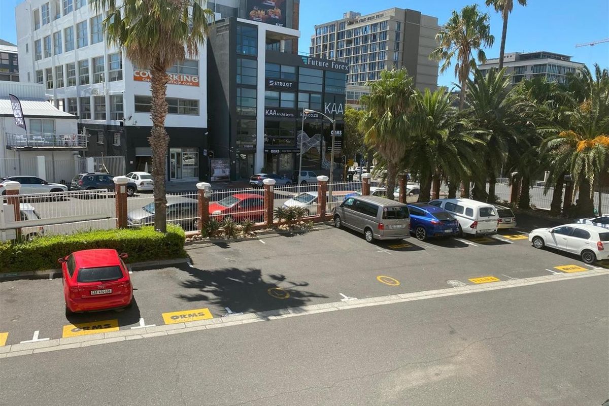 An elevated view of a paved commercial parking area featuring marked bays, perimeter fencing with brick pillars, and surrounding urban office buildings.