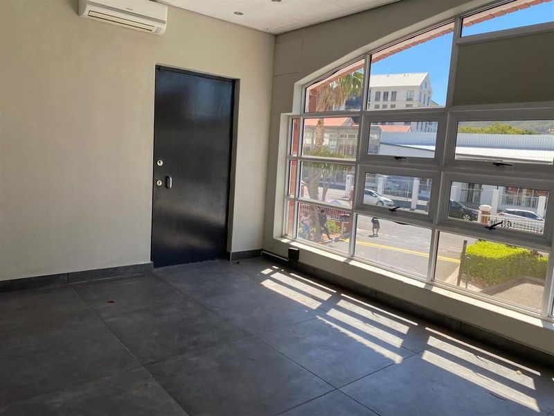A bright, open-plan retail space featuring large windows, tiled flooring, and a wall-mounted air conditioning unit, offering excellent natural light and street visibility.