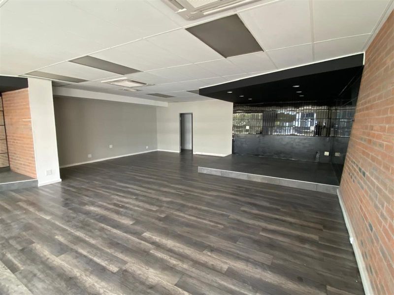 Spacious open-plan retail area featuring modern laminate flooring, exposed brick accents, a raised platform stage, and a suspended ceiling with recessed lighting.