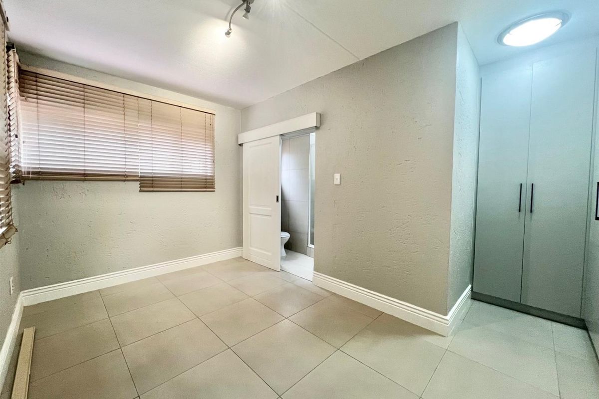 A bright bedroom featuring neutral tiled flooring, built-in cupboards, and an en-suite bathroom accessed via a sliding door.
