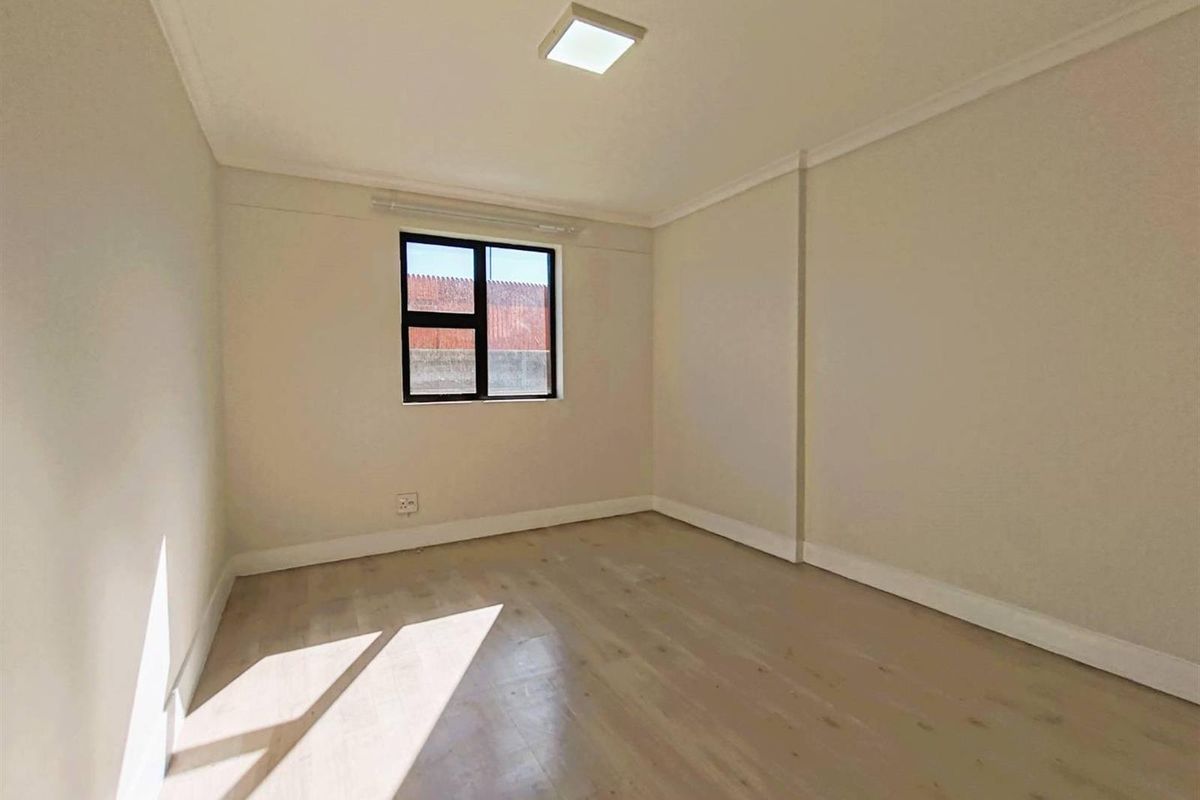 A bright, empty bedroom featuring light-toned laminate flooring, neutral walls, and a modern square ceiling light fixture.