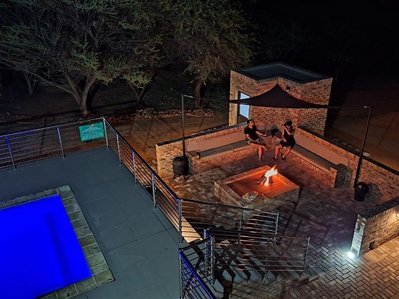 An elevated night view of a paved pool area featuring a built-in brick fire pit with integrated seating and stainless steel safety railings.