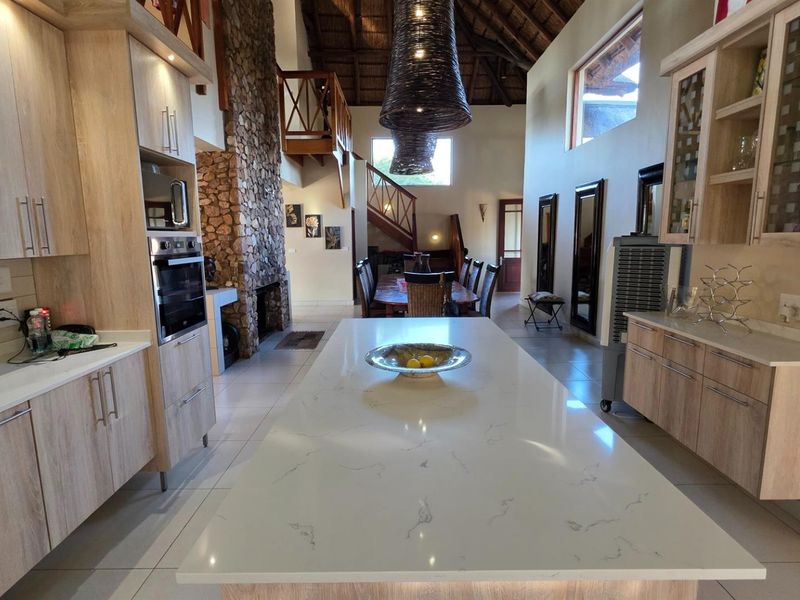Spacious open-plan kitchen and living area featuring a large stone-topped island, built-in oven, impressive stone fireplace, and a mezzanine level under a thatched roof.