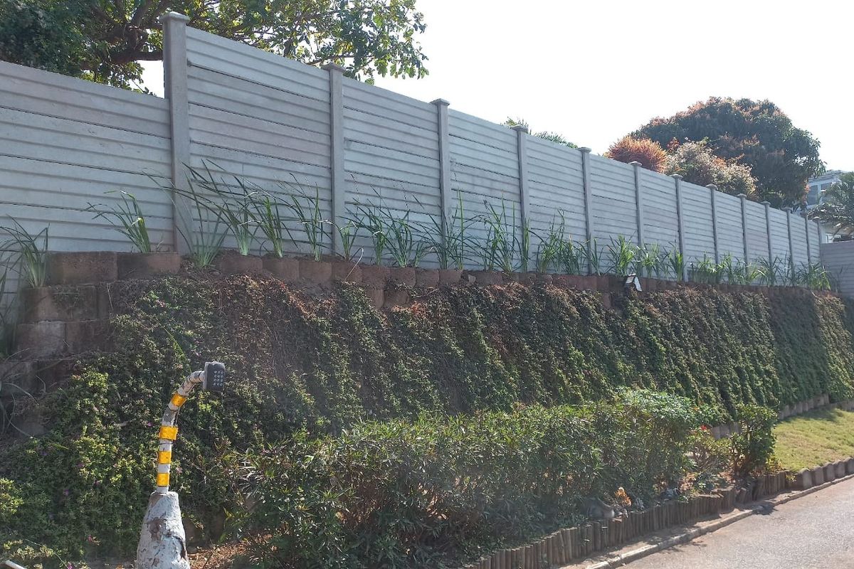 A view of a rear property boundary featuring a tall precast concrete wall set atop a landscaped retaining wall embankment.