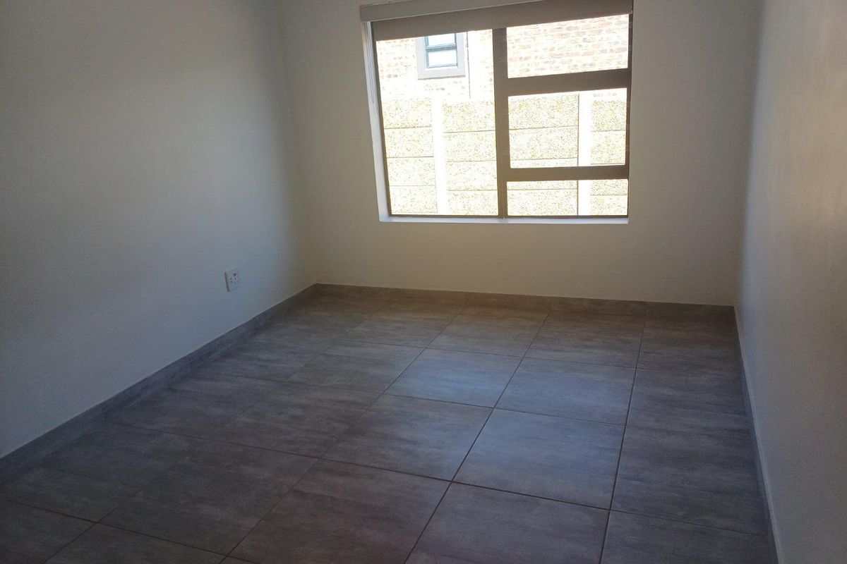 A clean, empty bedroom featuring neutral grey floor tiles, white walls, and a large window with a roller blind.