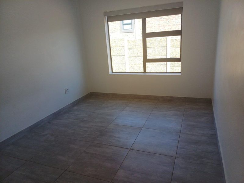 A clean, empty bedroom featuring neutral grey floor tiles, white walls, and a large window with a roller blind.