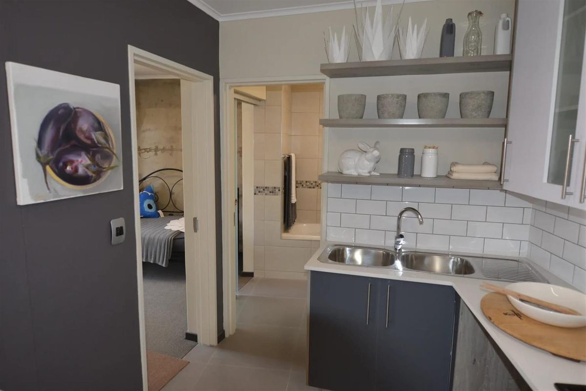 A compact kitchen area featuring modern cabinetry, a double stainless steel sink, subway tile backsplash, and open shelving, with views into an adjacent bedroom and bathroom.
