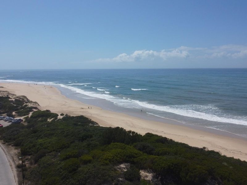 Aerial view of the coastline featuring a sandy beach, ocean waves, and adjacent coastal vegetation with a nearby road.