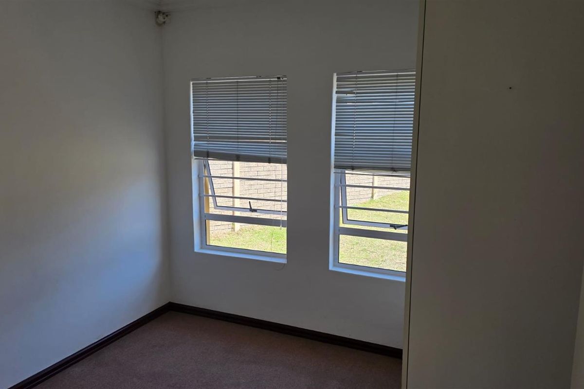 A simple bedroom featuring neutral walls, carpeted flooring, and two windows with fitted blinds overlooking the rear garden area.