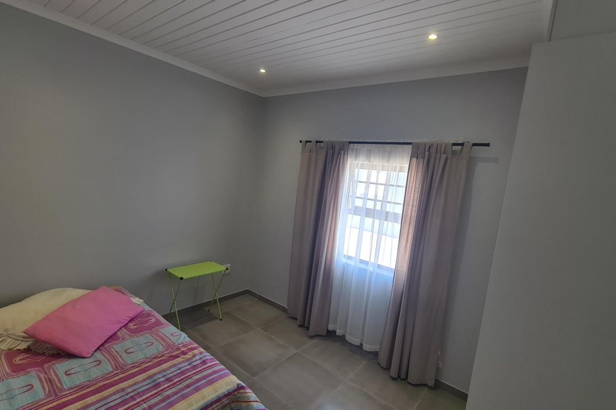 A clean, neutral-toned bedroom featuring tiled flooring, a white tongue-and-groove ceiling with recessed lighting, and a window with curtain fixtures.