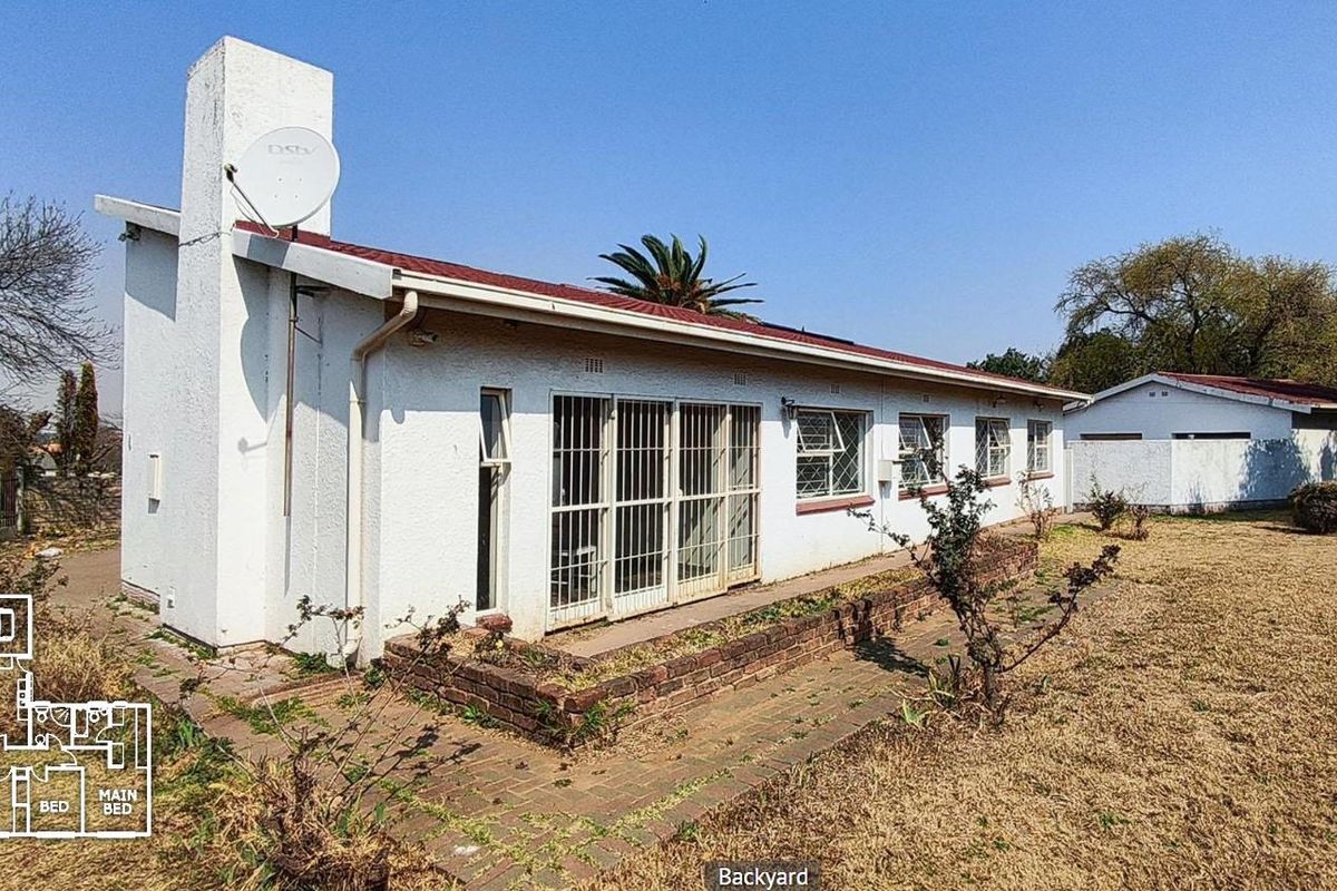 Rear view of the house featuring a brick chimney, metal roof, security-barred windows, and a detached garage set within a large, dry garden area.