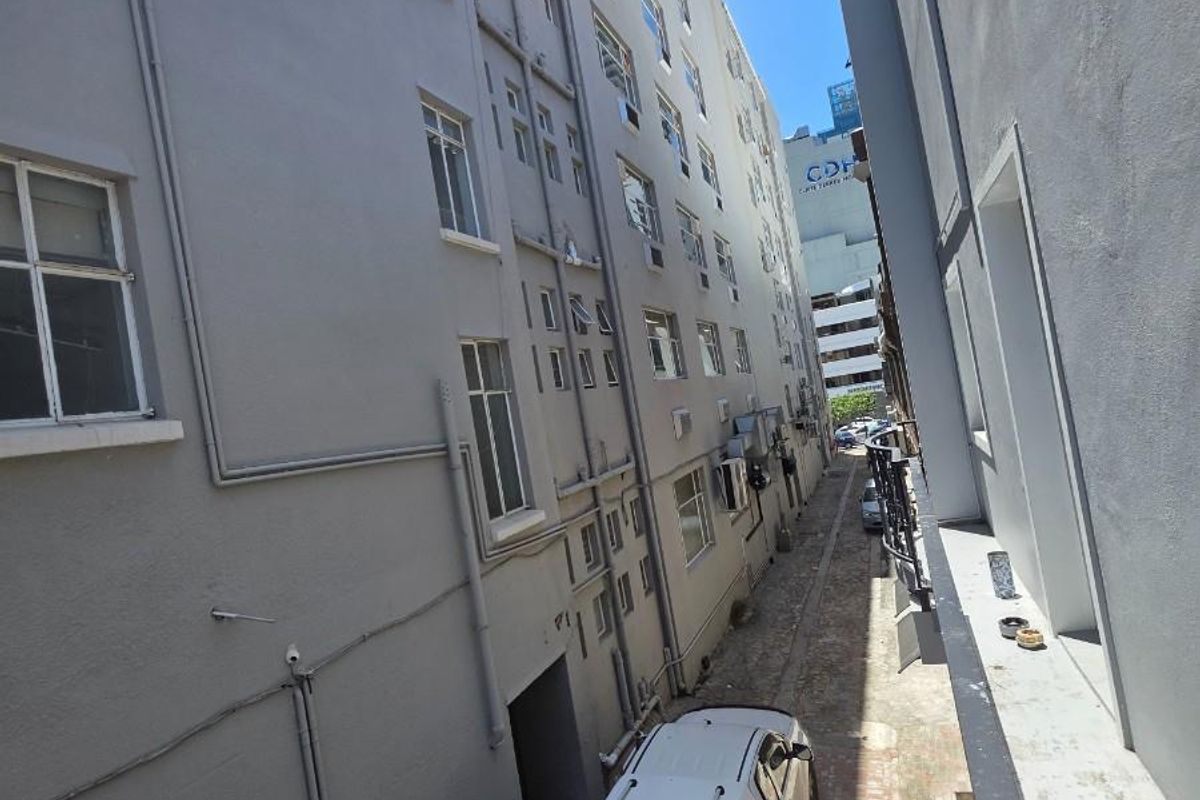 A view of the rear exterior alleyway between apartment buildings, featuring paved ground and multi-story facade with external window and plumbing infrastructure.