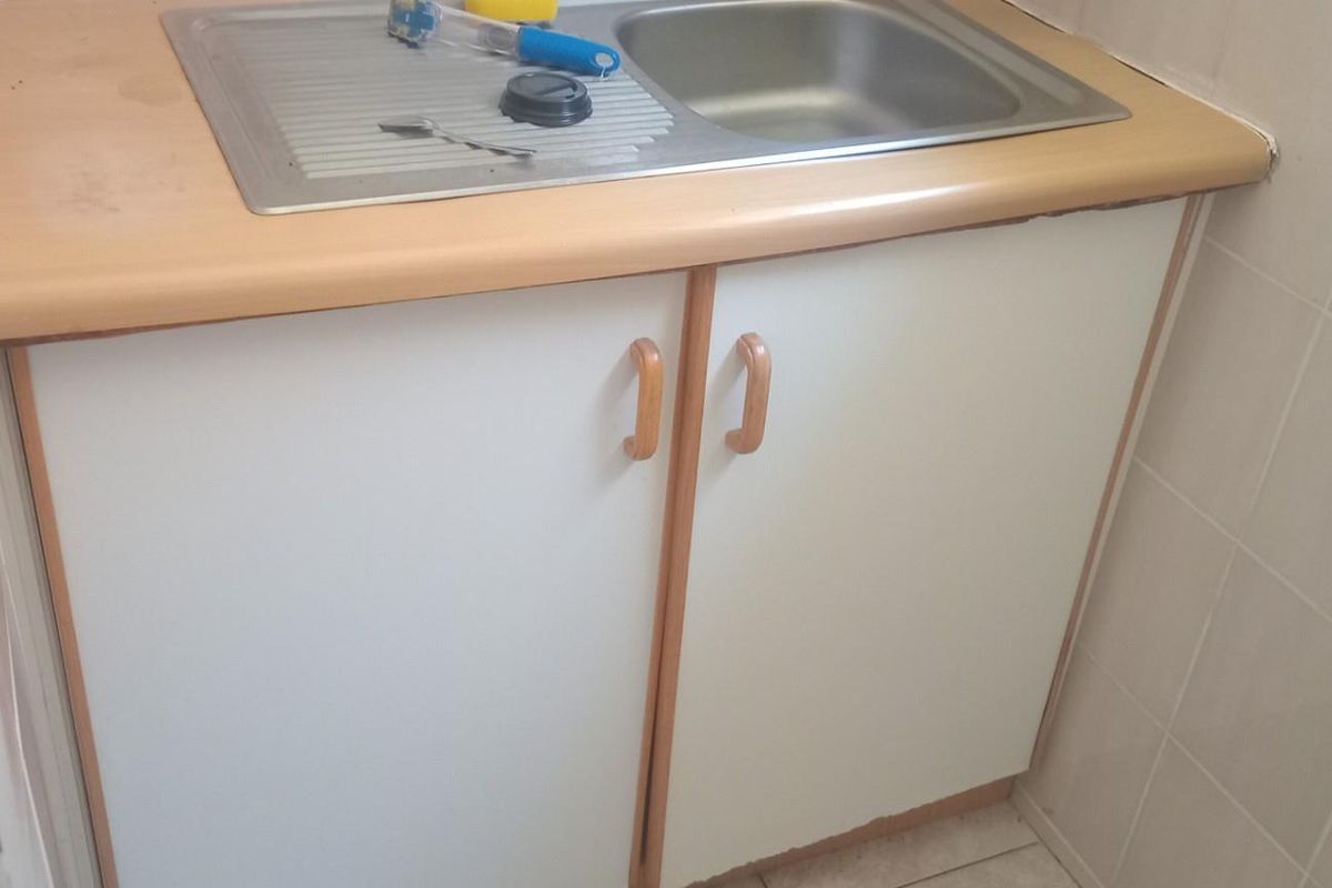A compact kitchenette area featuring a stainless steel sink, laminate countertop, and white base cabinetry with tiled wall finishes.