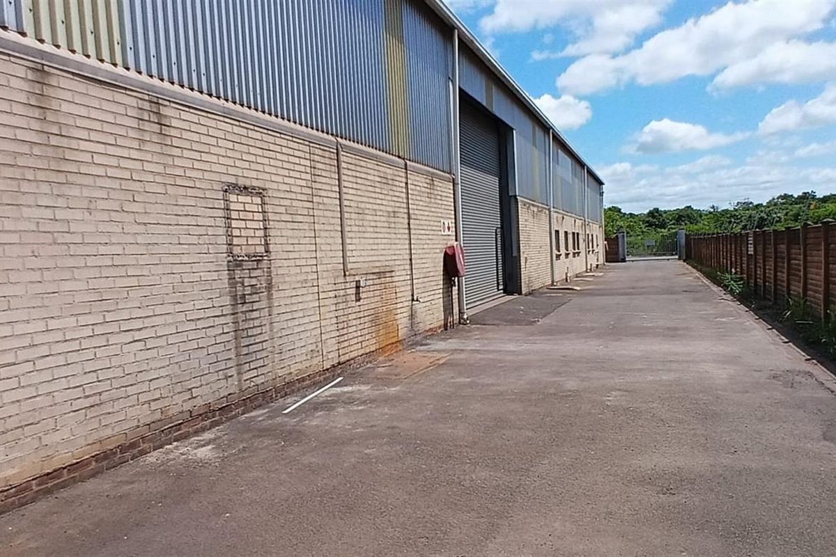 Side exterior view of an industrial warehouse featuring a brick facade, corrugated metal upper cladding, roller shutter door, and concrete driveway.