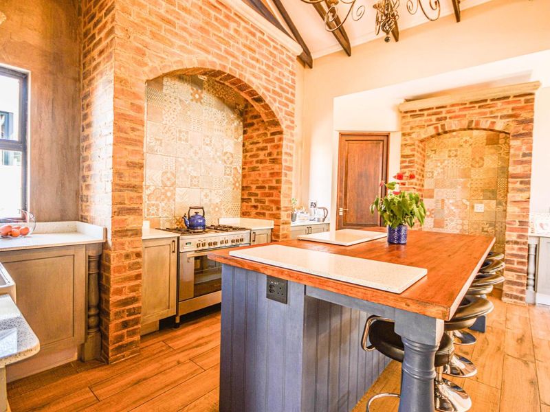A rustic-style kitchen featuring exposed brick archways, a large timber-topped island, gas stove, and decorative tiled splashbacks under exposed ceiling beams.