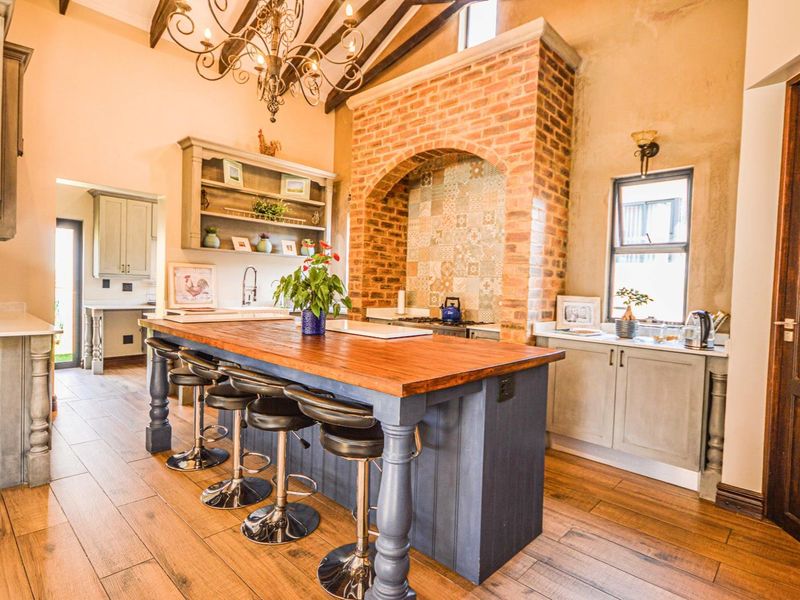 A rustic-style kitchen featuring a large timber-topped island, exposed vaulted ceiling beams, and a distinctive brick feature wall with decorative tiling.