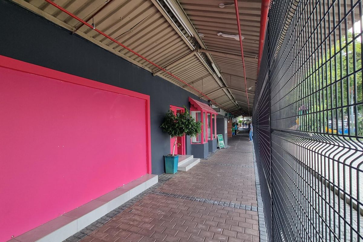 A covered outdoor walkway featuring a paved path, metal security fencing, and retail storefronts with painted facade details.