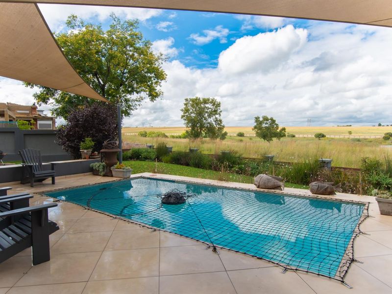 Outdoor pool area featuring a tiled patio, shade sail coverage, and a safety net, overlooking an expansive open field landscape.