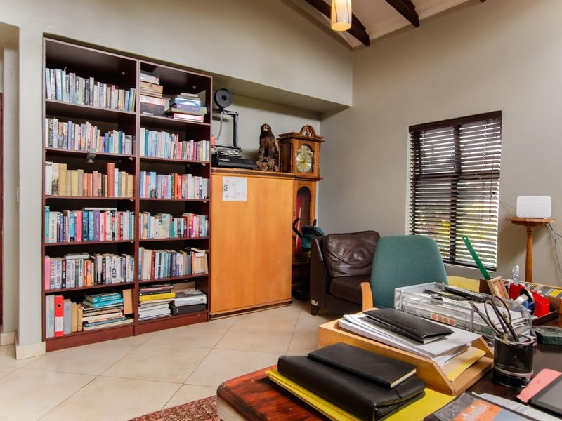 A functional home office featuring built-in floor-to-ceiling shelving, exposed timber ceiling beams, and tiled flooring with natural light through venetian blinds.