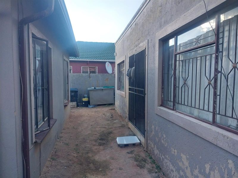 A narrow side passage between two residential structures featuring security gates, metal window bars, and an unpaved ground surface.