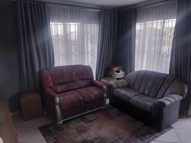 A living room featuring tiled flooring, large windows with security bars, and floor-to-ceiling curtains, providing a functional and well-lit space.