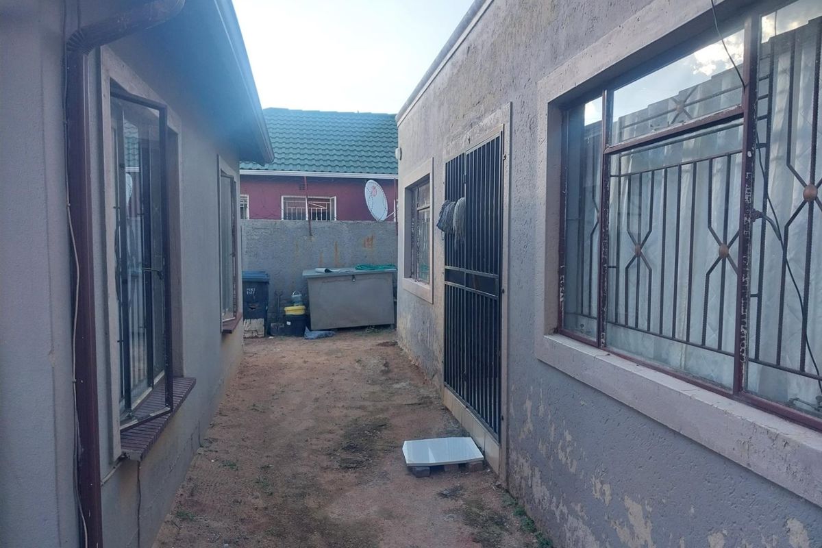 A narrow side passage between two residential structures featuring security gates, metal window bars, and an unpaved ground surface.