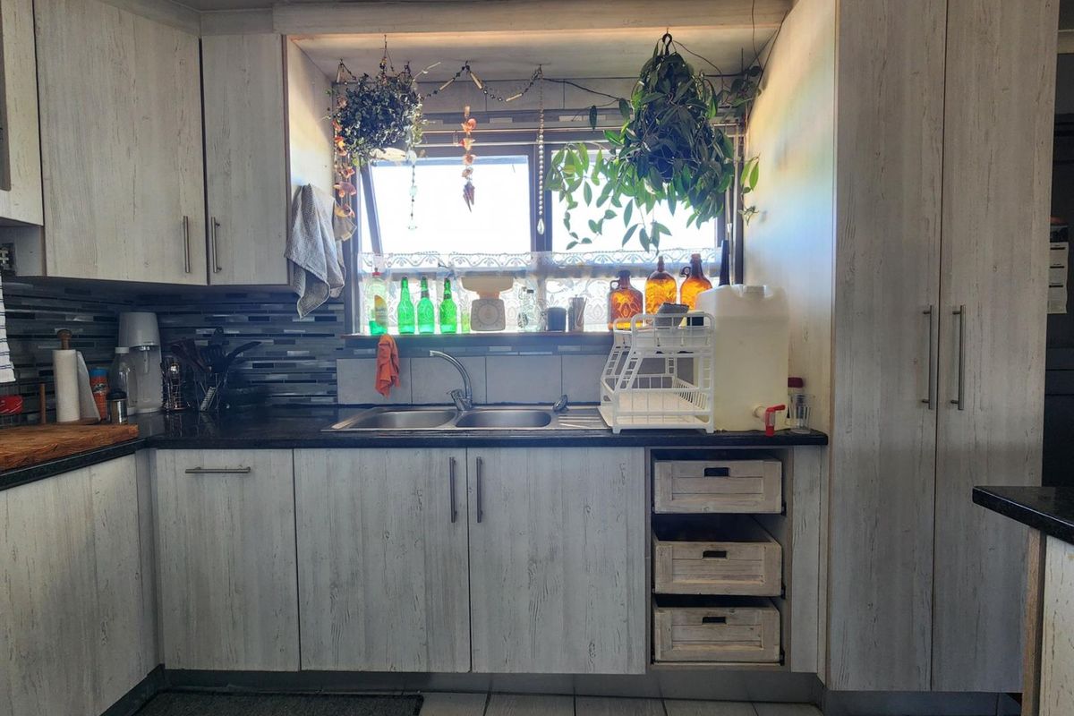 A functional kitchen featuring light wood-grain cabinetry, a double-basin sink, dark countertops, and a tiled backsplash with natural light from the window.